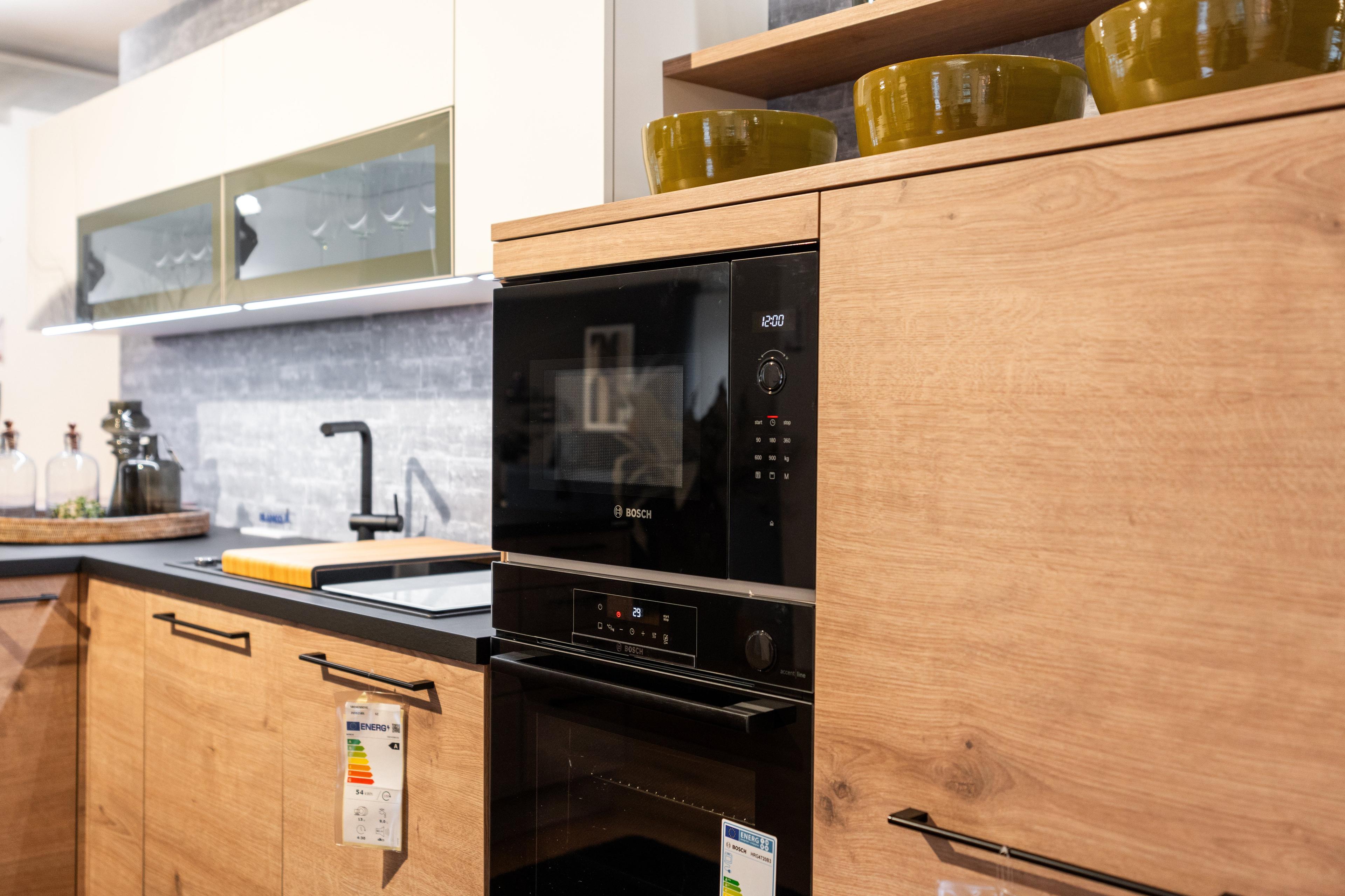 Nahaufnahme der eingebauten schwarzen Geräte - Mikrowelle und Backofen - in hellen Holzfronten. Offene Regale mit gelben Schalen, Oberschränke mit Glasfronten und gelben Akzenten.