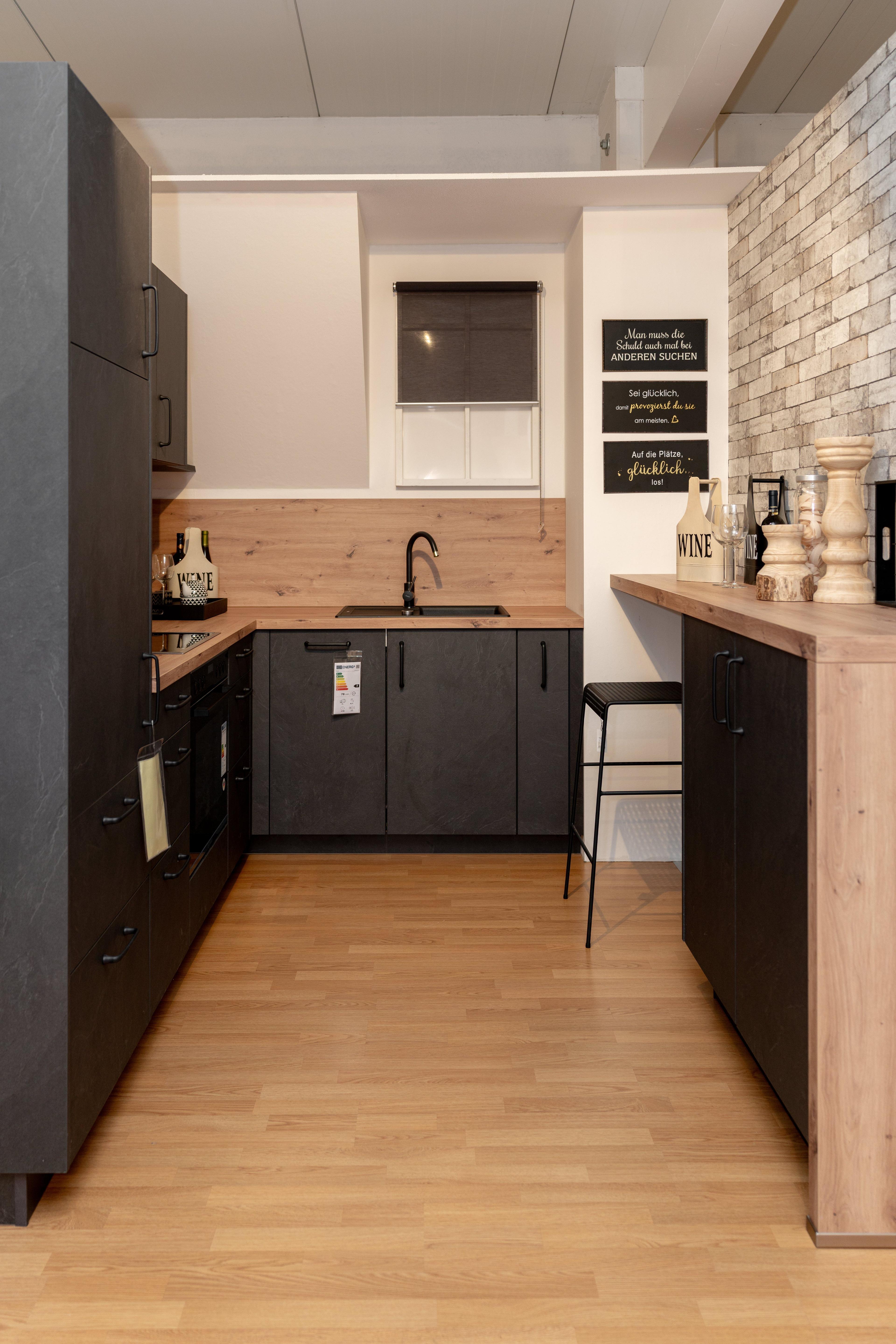 Moderne Küche im Industrial-Style mit anthrazitfarbenen Schränken, Holzarbeitsplatte und -rückwand. Backsteinwand rechts, deutsche Sprüche an der Wand und schwarzer Barhocker vor der Kücheninsel.
