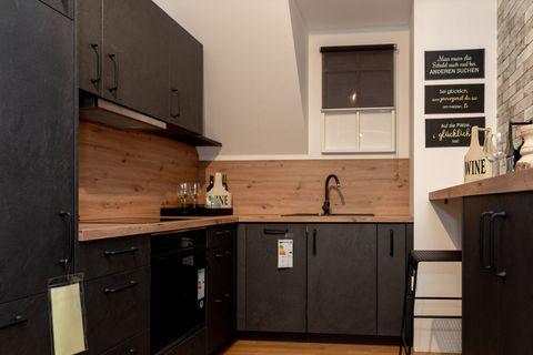 Moderne Küche im Industrial-Style mit anthrazitfarbenen Schränken, Holzarbeitsplatte und -rückwand. Backsteinwand rechts, deutsche Sprüche an der Wand und schwarzer Barhocker vor der Kücheninsel.