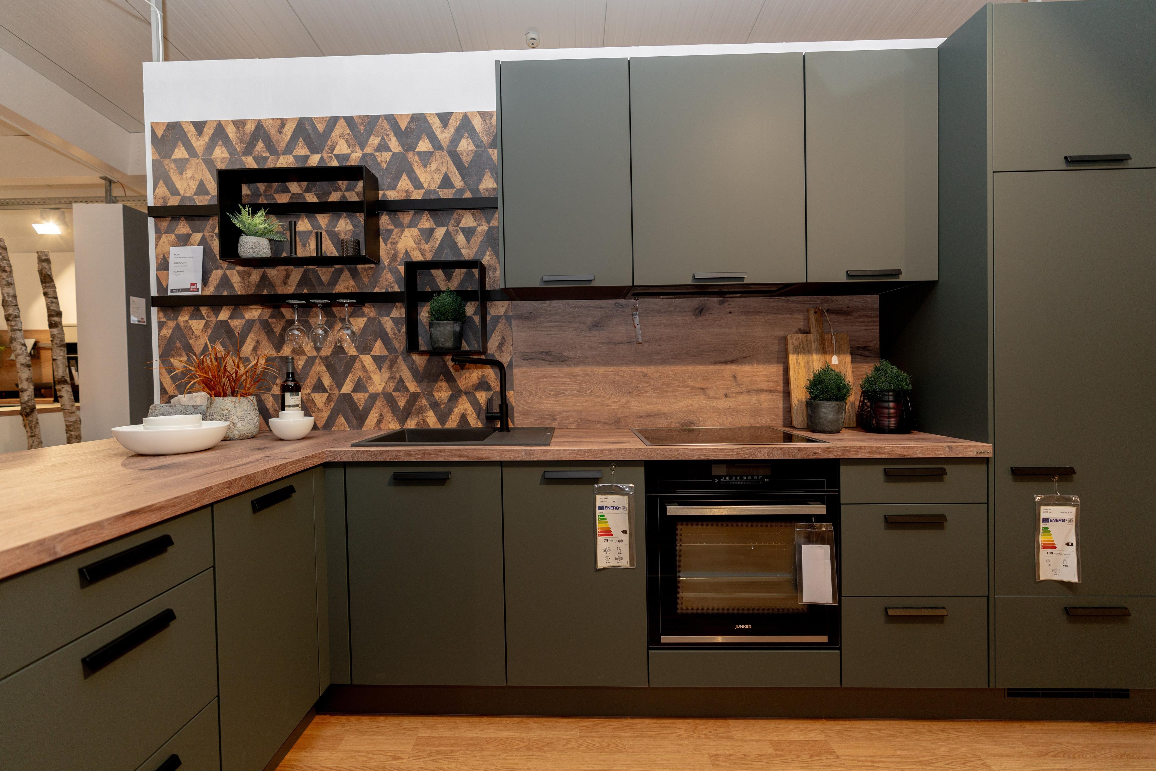 Detailansicht der olivgrünen Küche mit geometrischer Dreiecks-Wandfliese in Holzoptik, schwarzen schwebenden Regalen und Holzrückwand. Eingebauter Backofen und Dekoration mit Pflanzen.