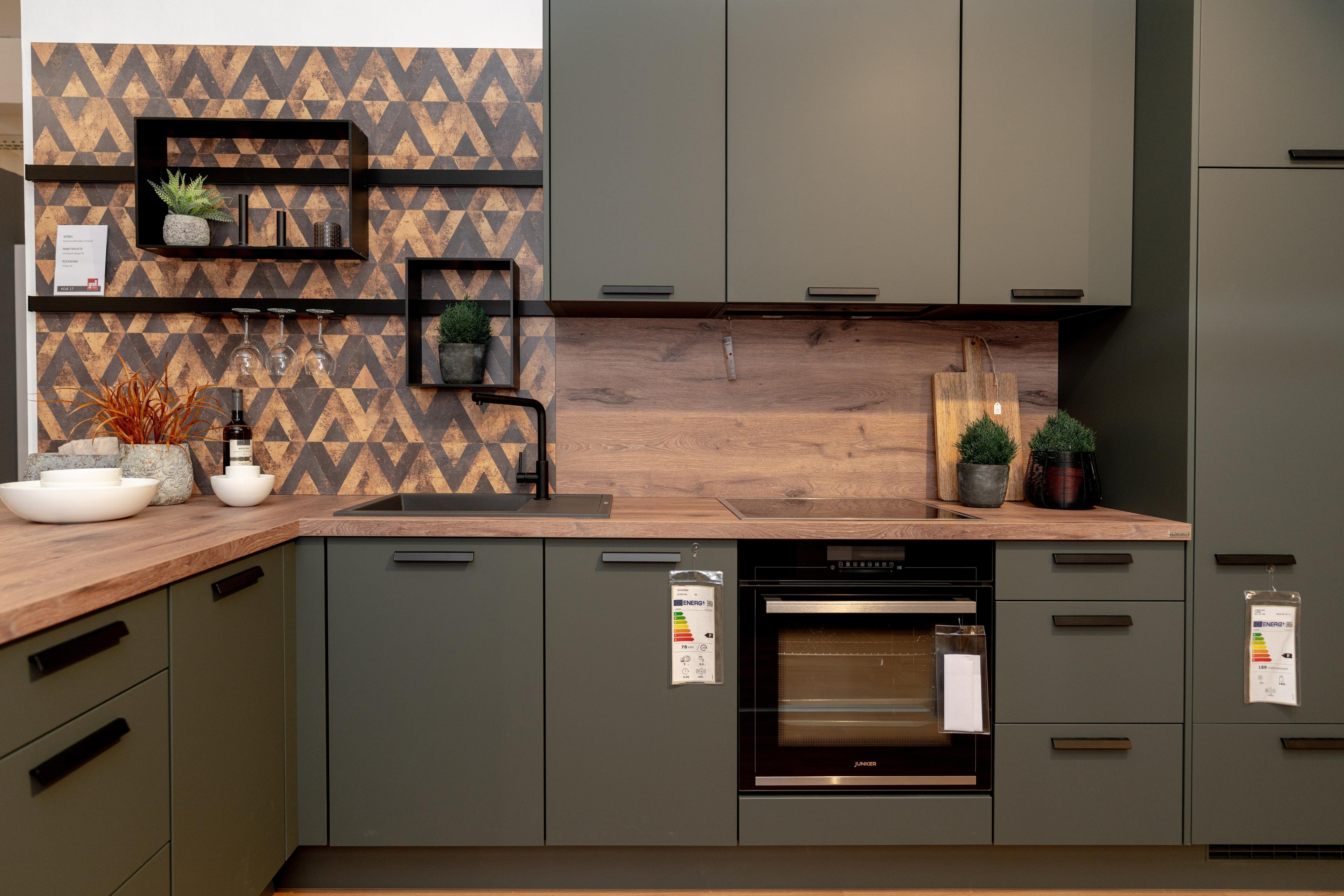 Frontalansicht der Küchenzeile mit olivgrünen Schränken, Holzarbeitsplatte und geometrischen Wandfliesen. Schwarze Metallregale mit Gläsern und Pflanzen, eingebauter Backofen in der Mitte.