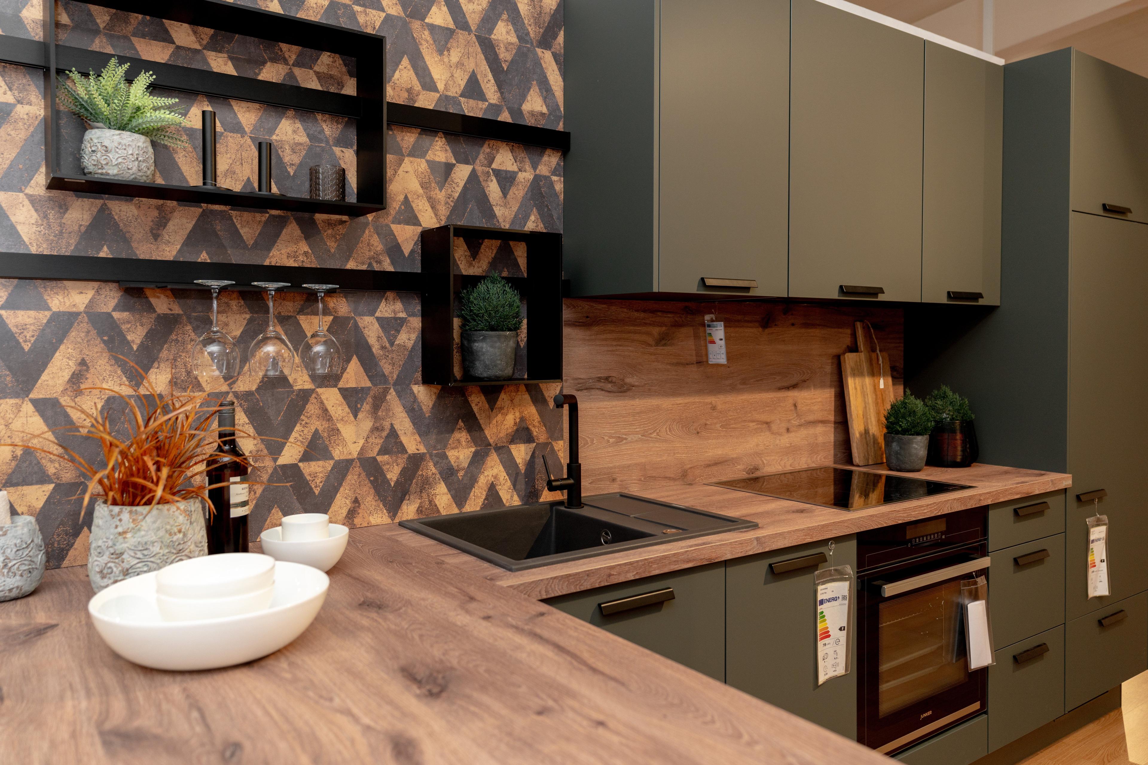Nahaufnahme der Arbeitsplatte mit weißen Keramikschalen, schwarzer Spüle und geometrischer Wandfliese. Schwarze schwebende Regale mit Pflanzen und Weingläsern, natürliche Holzmaserung der Arbeitsplatte.