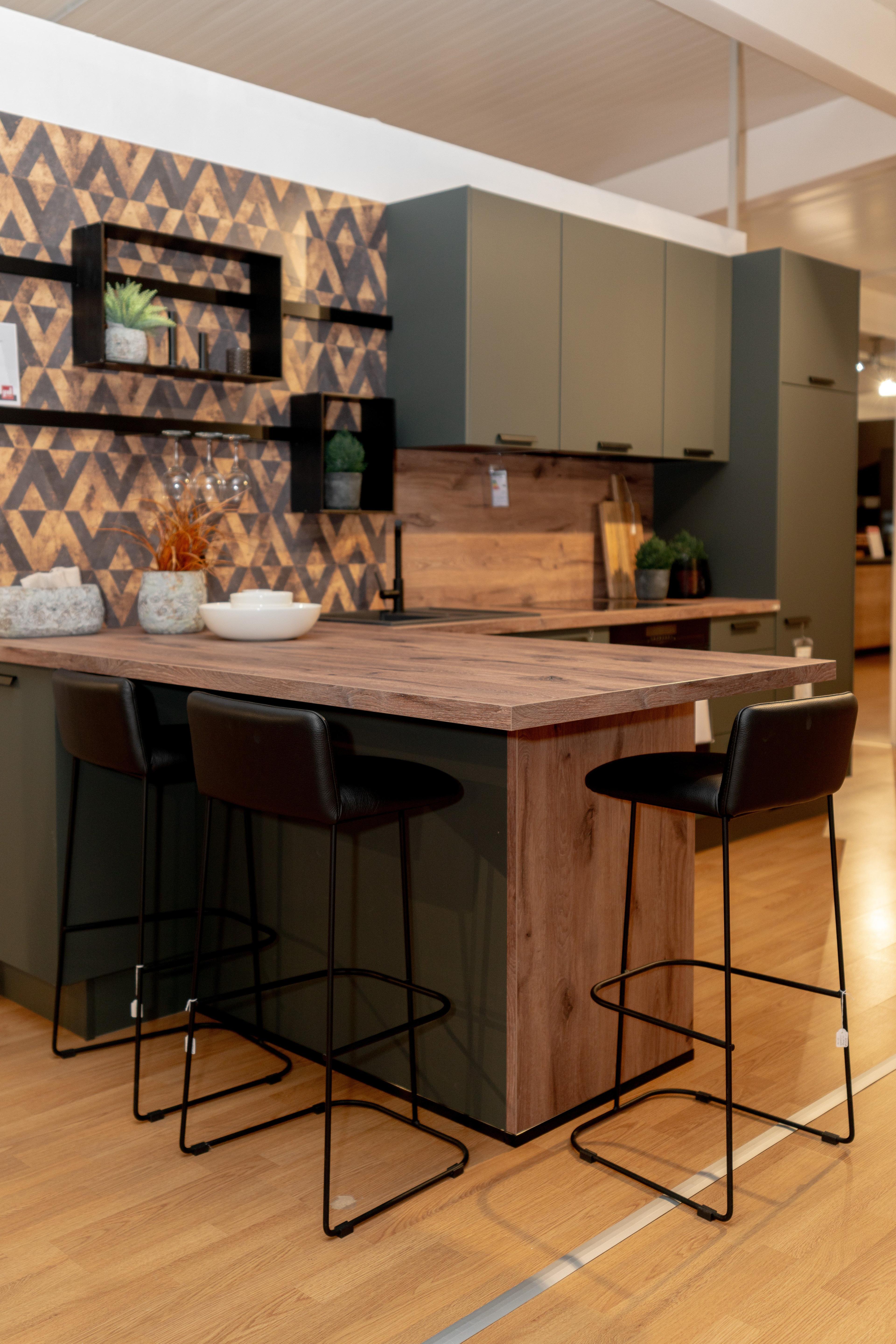 Kücheninsel aus Holz mit vier schwarzen Barhockern, olivgrüne Küchenschränke im Hintergrund. Geometrische Dreiecks-Wandfliese und schwarze schwebende Regale mit Dekoration.