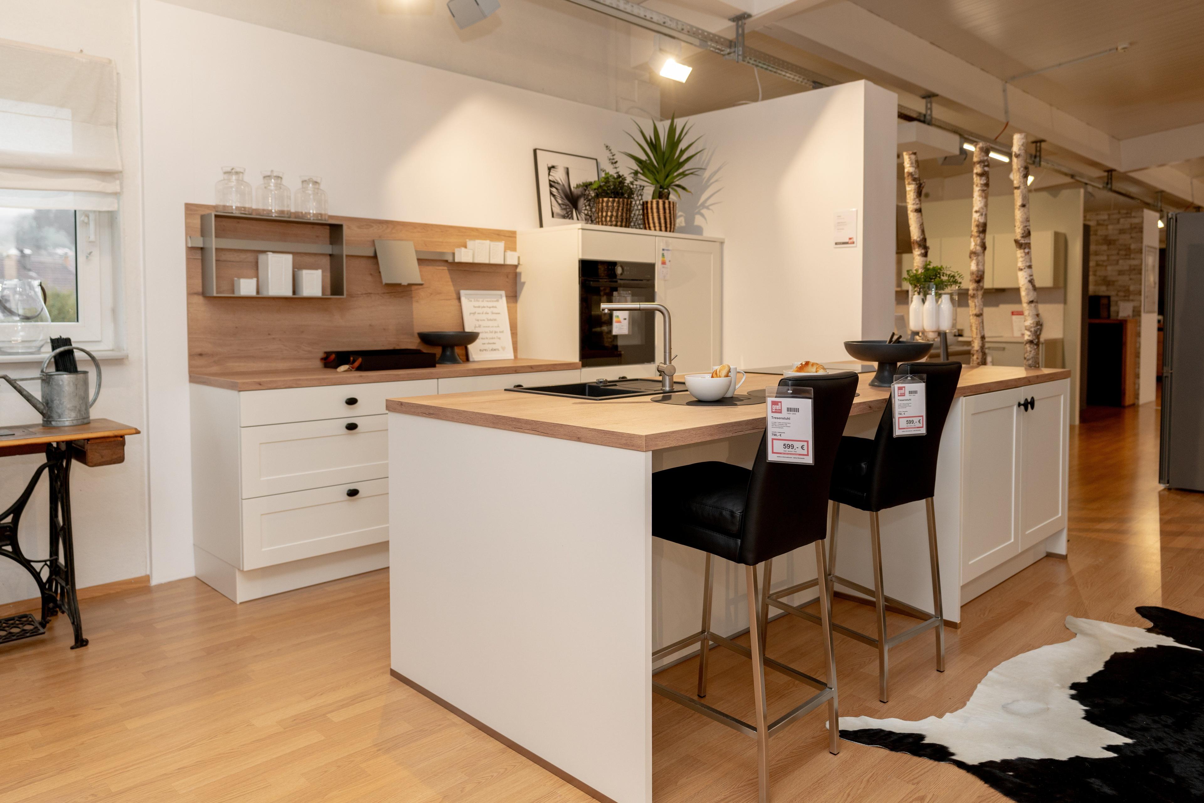 Moderne weiße Küche im Landhausstil mit Holzarbeitsplatte, Kücheninsel mit zwei schwarzen Barhockern. Offene Holzregale mit Dekoration, Showroom-Ambiente mit Birkenstämmen und schwarz-weißem Kuhfell-Teppich.