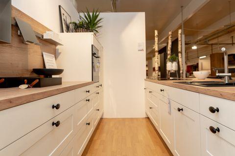 Moderne weiße Küche im Landhausstil mit Holzarbeitsplatte, Kücheninsel mit zwei schwarzen Barhockern. Offene Holzregale mit Dekoration, Showroom-Ambiente mit Birkenstämmen und schwarz-weißem Kuhfell-Teppich.