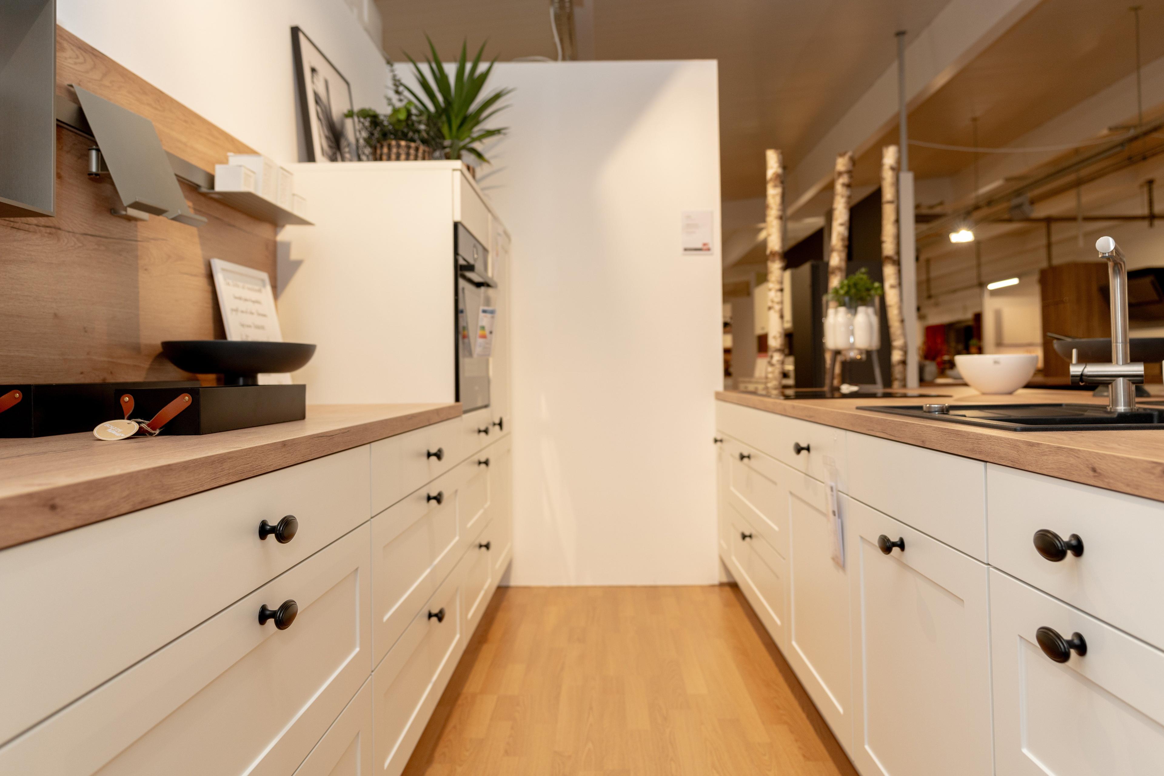 Schmale Küchenzeile mit weißen Kassettenfronten und schwarzen Knöpfen, beidseitig angeordnet mit Gang in der Mitte. Holzarbeitsplatten, eingebaute Mikrowelle und Showroom-Beleuchtung an der Decke.