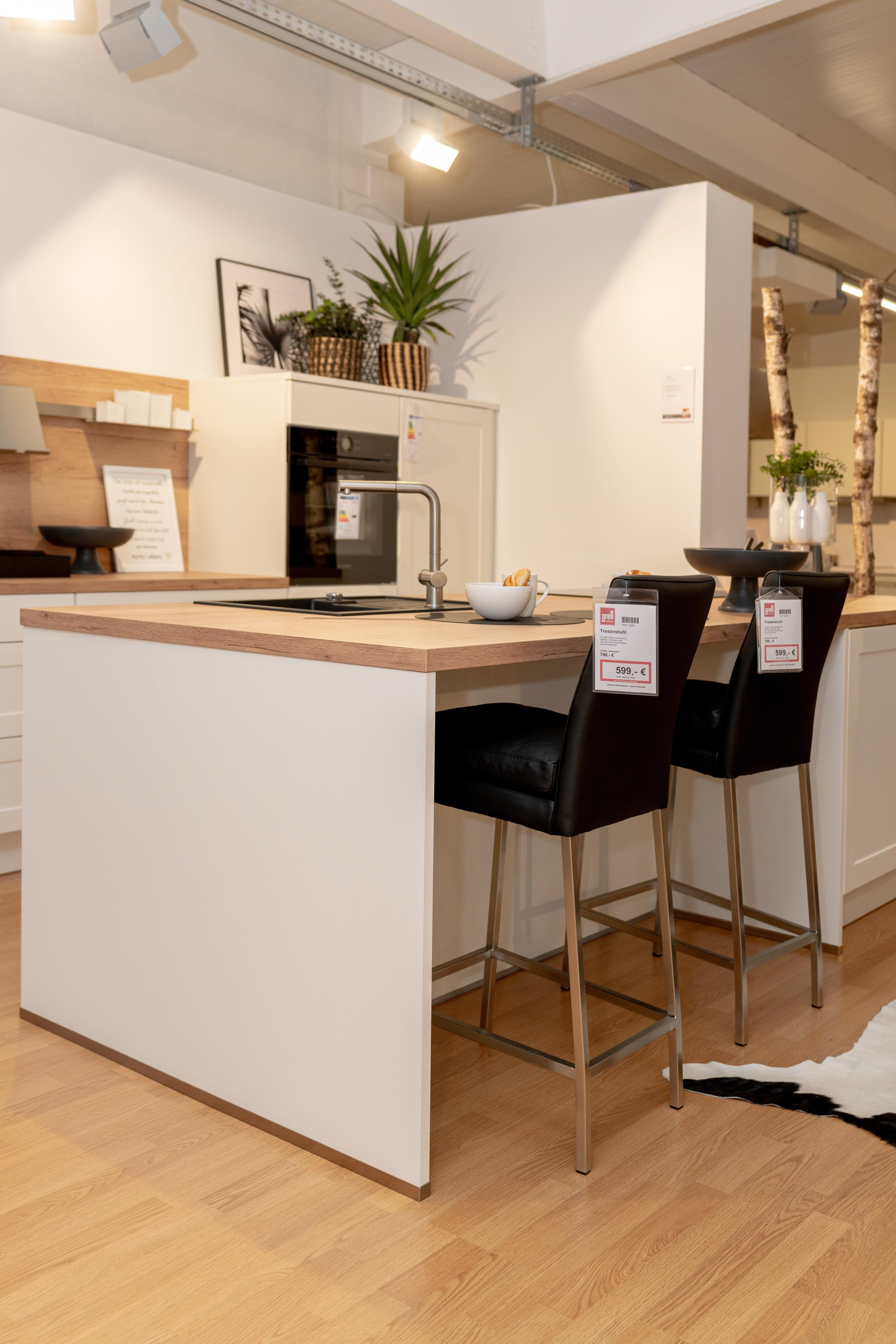 Seitliche Ansicht der Kücheninsel mit eingebauter Spüle, Chromarmatur und zwei schwarzen Barhockern. Weiße Schränke im Landhausstil, Holzarbeitsplatte und Showroom-Atmosphäre mit industrieller Deckenbeleuchtung.