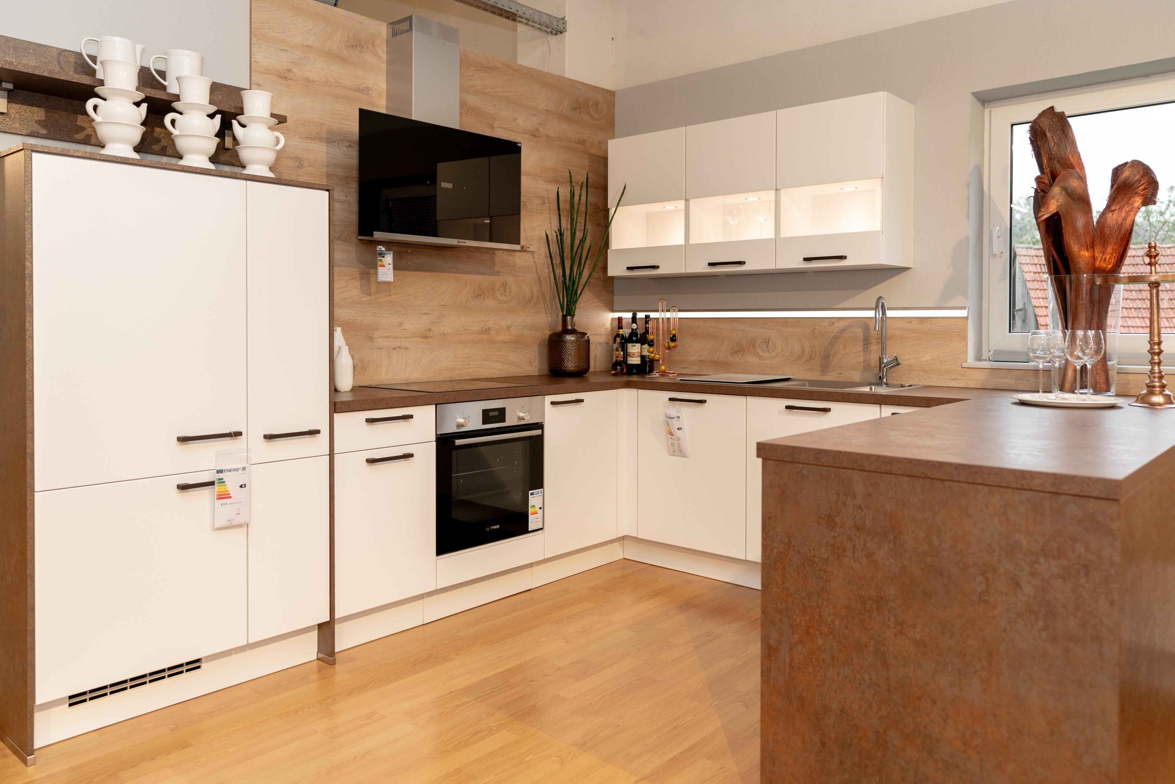 L-förmige Küche mit weißen Fronten und Holzarbeitsplatte, dunkelbraune Kücheninsel im Vordergrund. Holzrückwand, eingebauter Backofen und Dekoration mit Pflanzen und Weinflaschen.