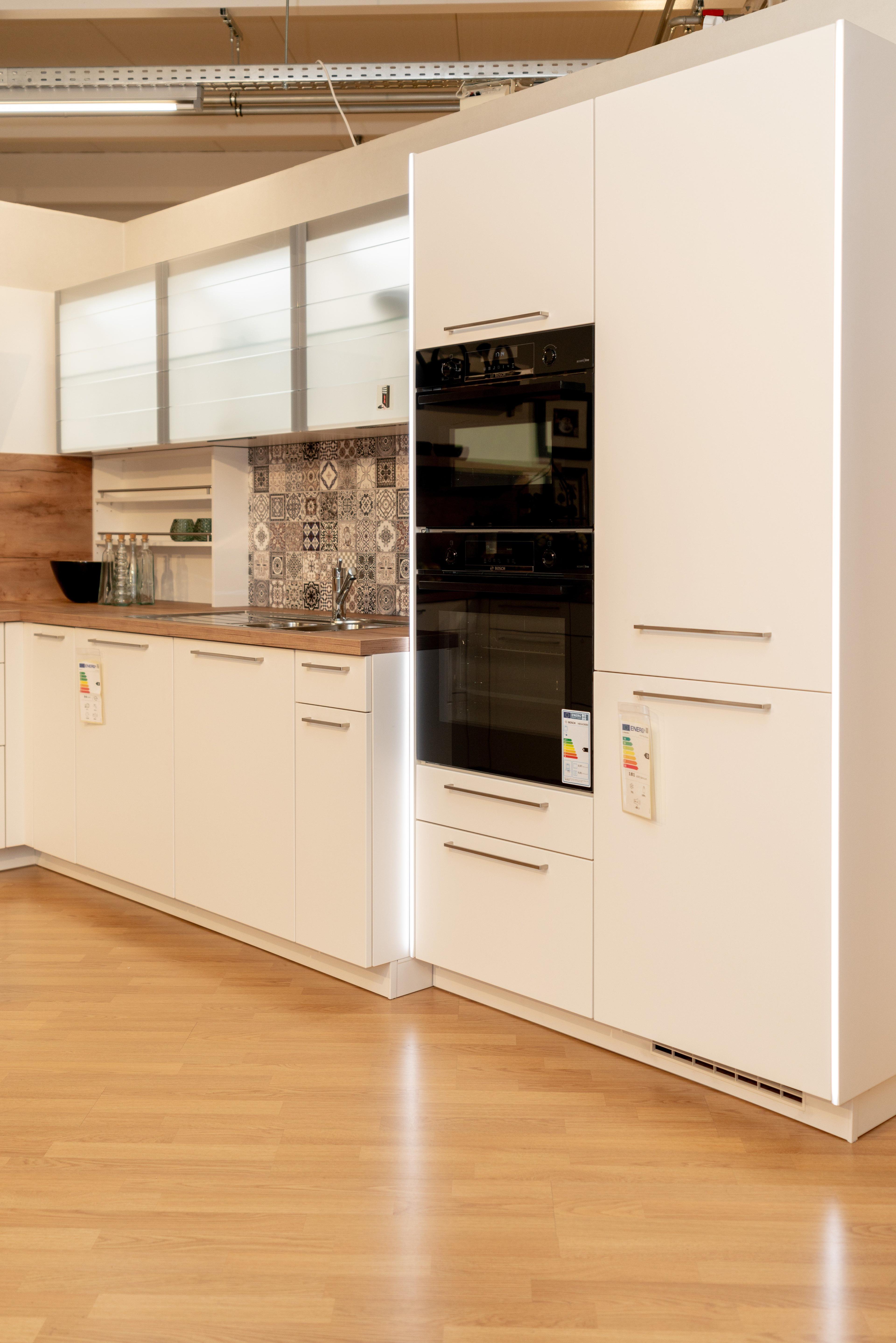 Seitliche Ansicht der weißen Küche mit eingebautem schwarzem Backofen, buntem Fliesenspiegel im Patchwork-Design und Holzarbeitsplatte. Showroom-Ambiente mit industrieller Decke.