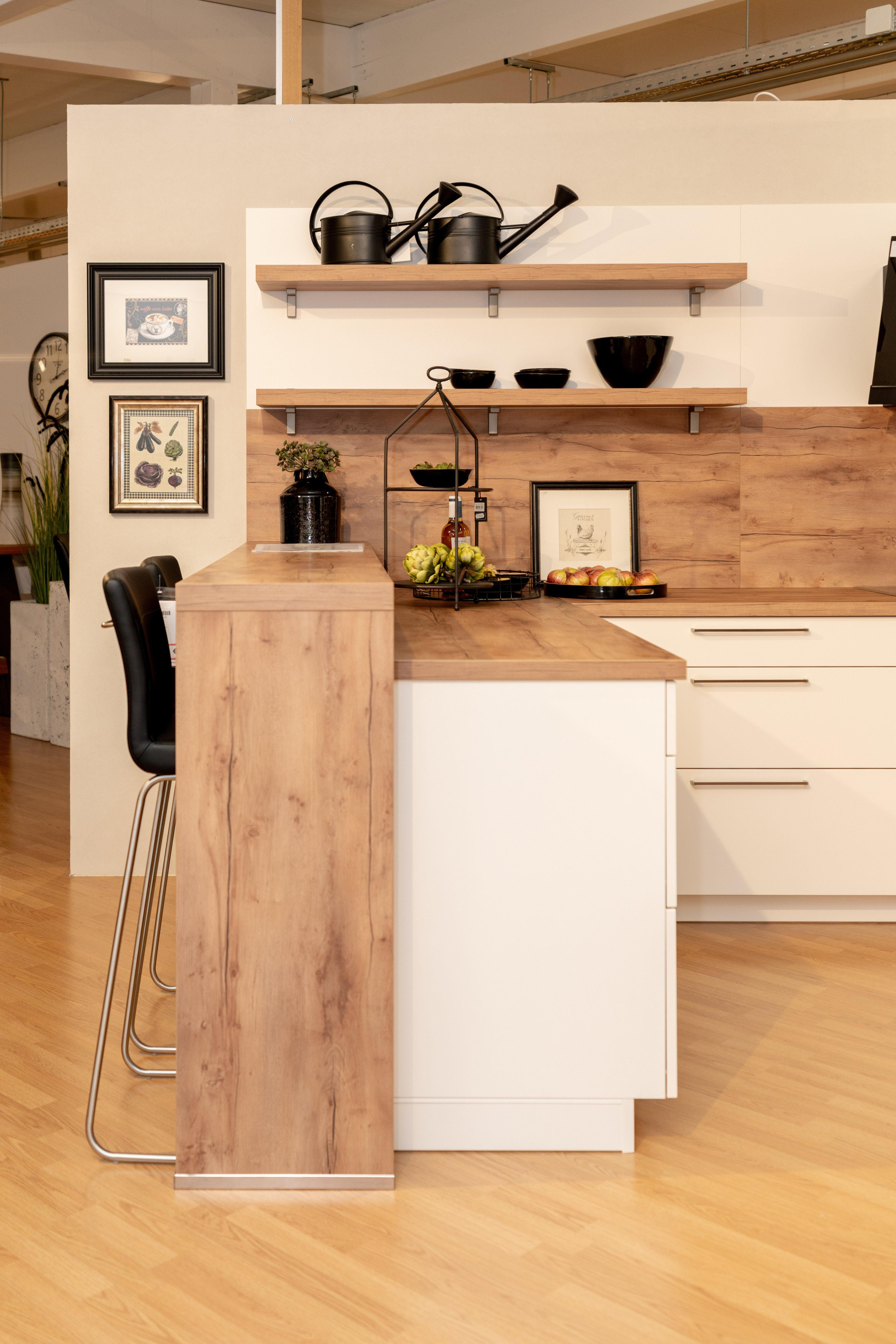 Detailansicht der Kücheninsel mit Holzverkleidung und weißer Front, schwarzer Barhocker davor. Holzregale mit schwarzen Schalen, Pflanzen und Dekoration, Holzrückwand mit natürlicher Maserung.