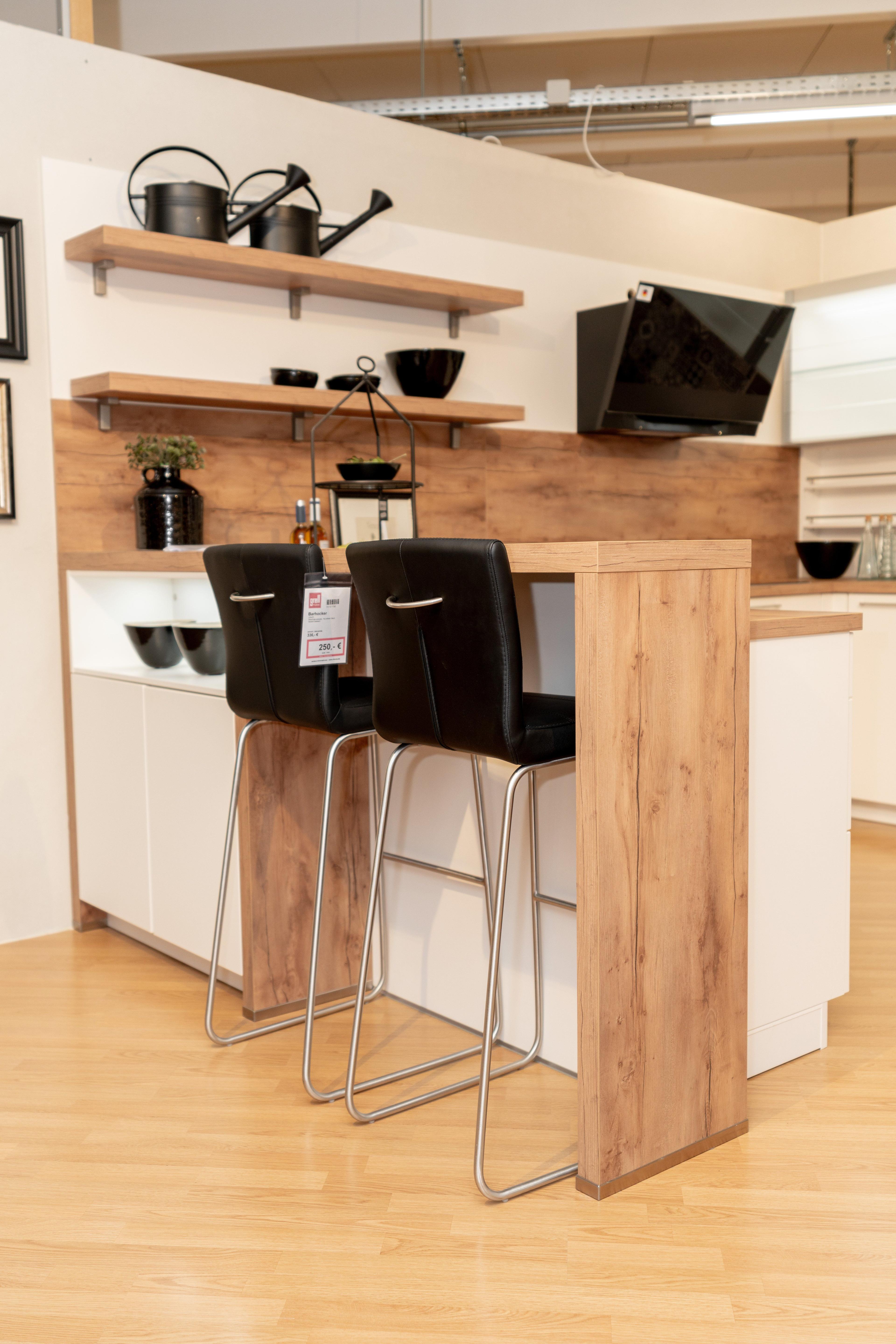 Nahaufnahme der Kücheninsel mit zwei schwarzen Barhockern mit Chromgestell und Holzsockel. Weiße Unterschränke, Holzregale mit schwarzem Geschirr und Gießkannen im Hintergrund.