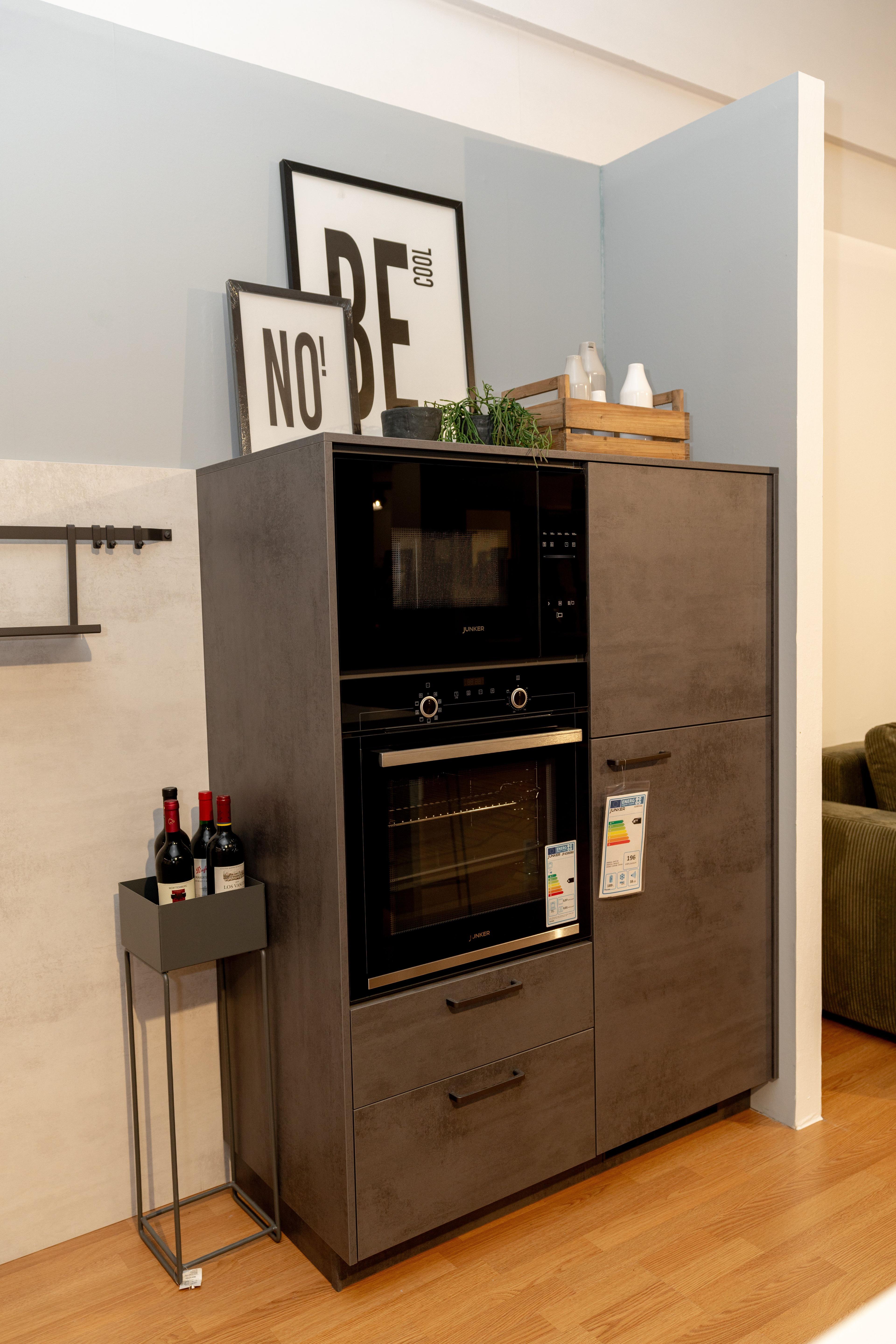 Hochschrank in Betonoptik mit eingebautem Backofen und Mikrowelle, gerahmte Poster "NO!" und "BE COOL" an der Wand. Schwarzer Beistellwagen mit Weinflaschen daneben.