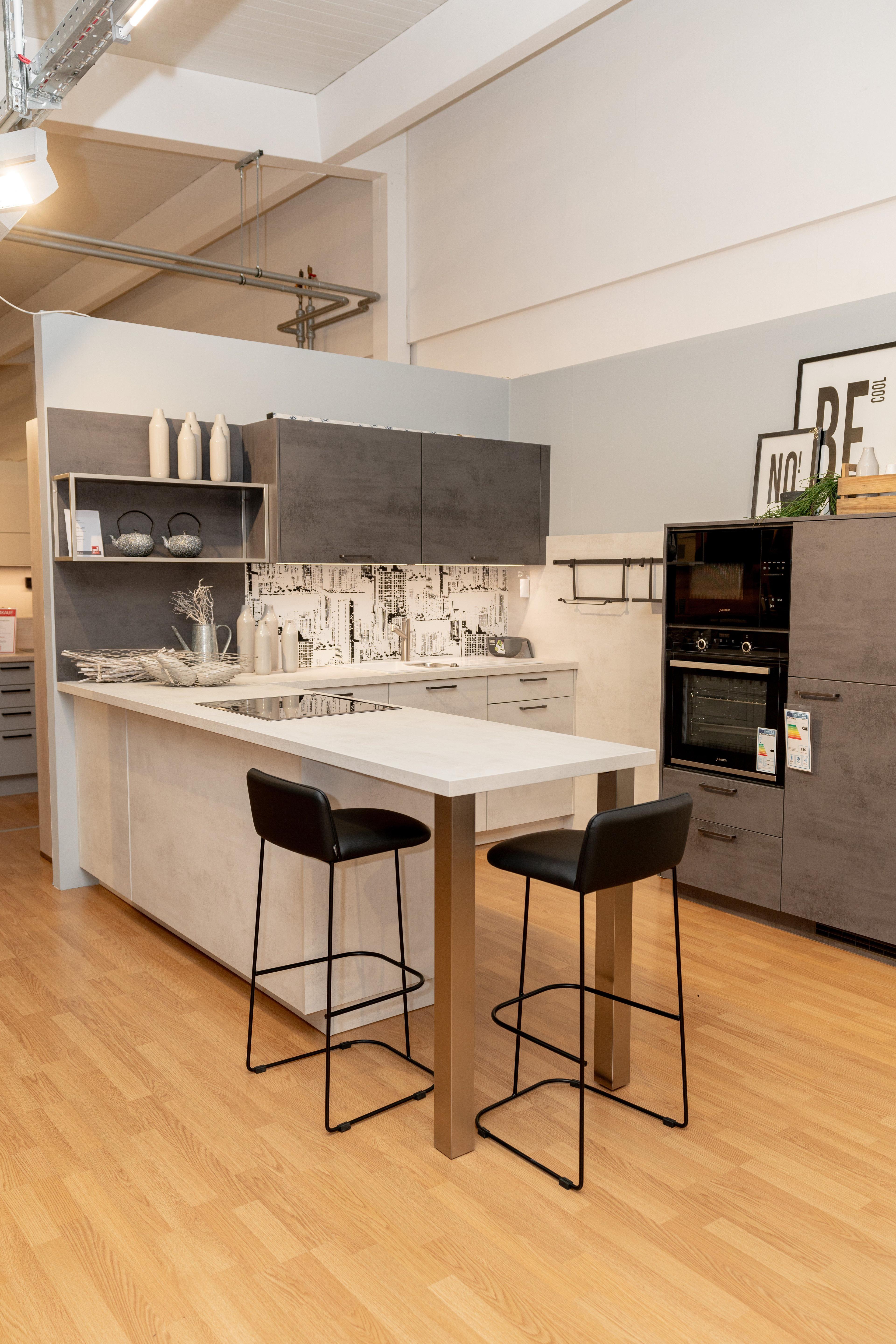 Gesamtansicht der Küche mit weißer Kücheninsel, zwei schwarzen Barhockern und anthrazitfarbenen Schränken. Stadtmotiv-Fliesenspiegel, offene Regale und Poster im Hintergrund, Showroom-Atmosphäre mit industrieller Decke.