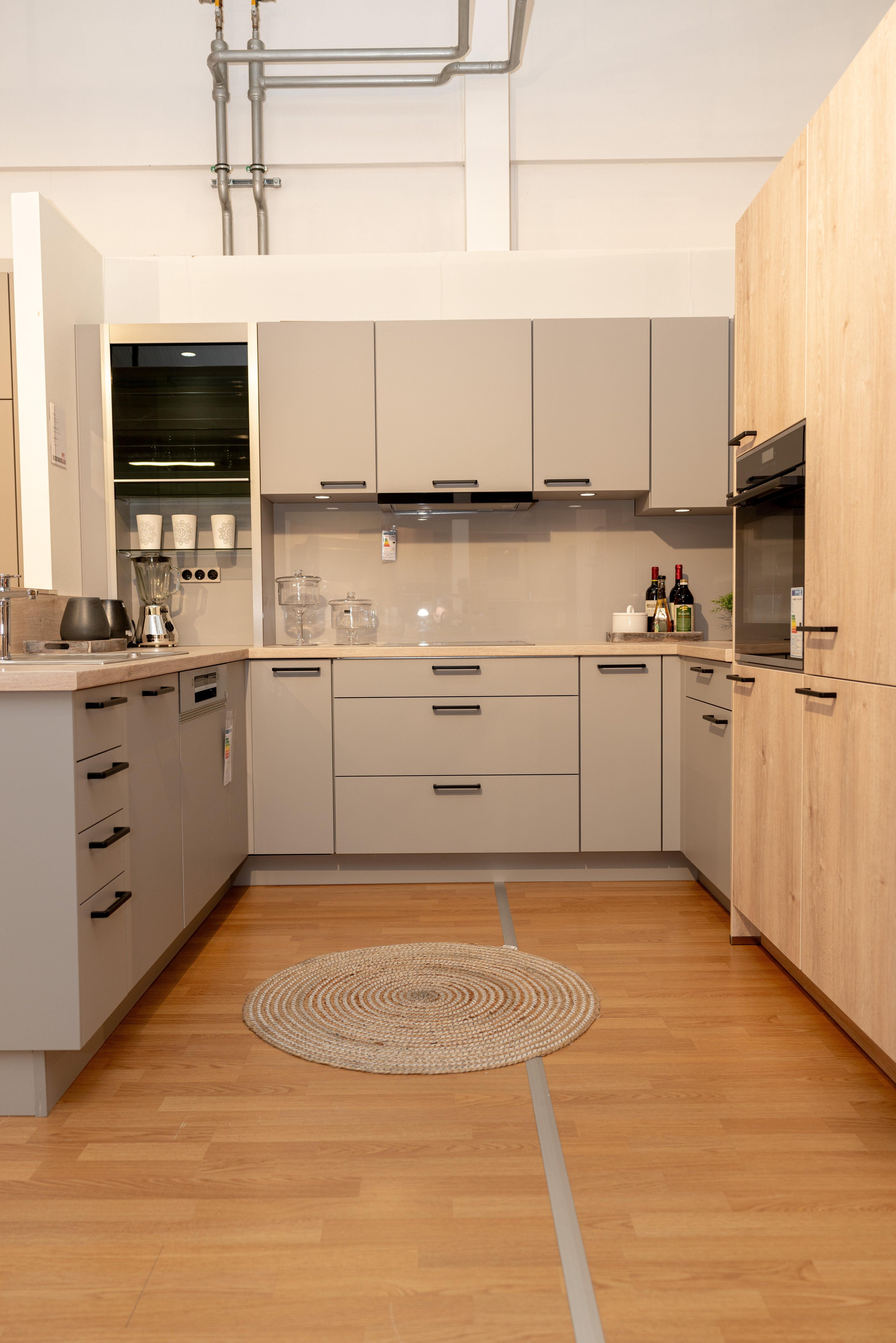 Frontale Ansicht der beigen U-Küche mit Holzarbeitsplatte, offenen Regalen mit Gläsern und Weinflaschen im Hintergrund. Geflochtener Rundteppich und Parkett, Showroom-Ambiente.