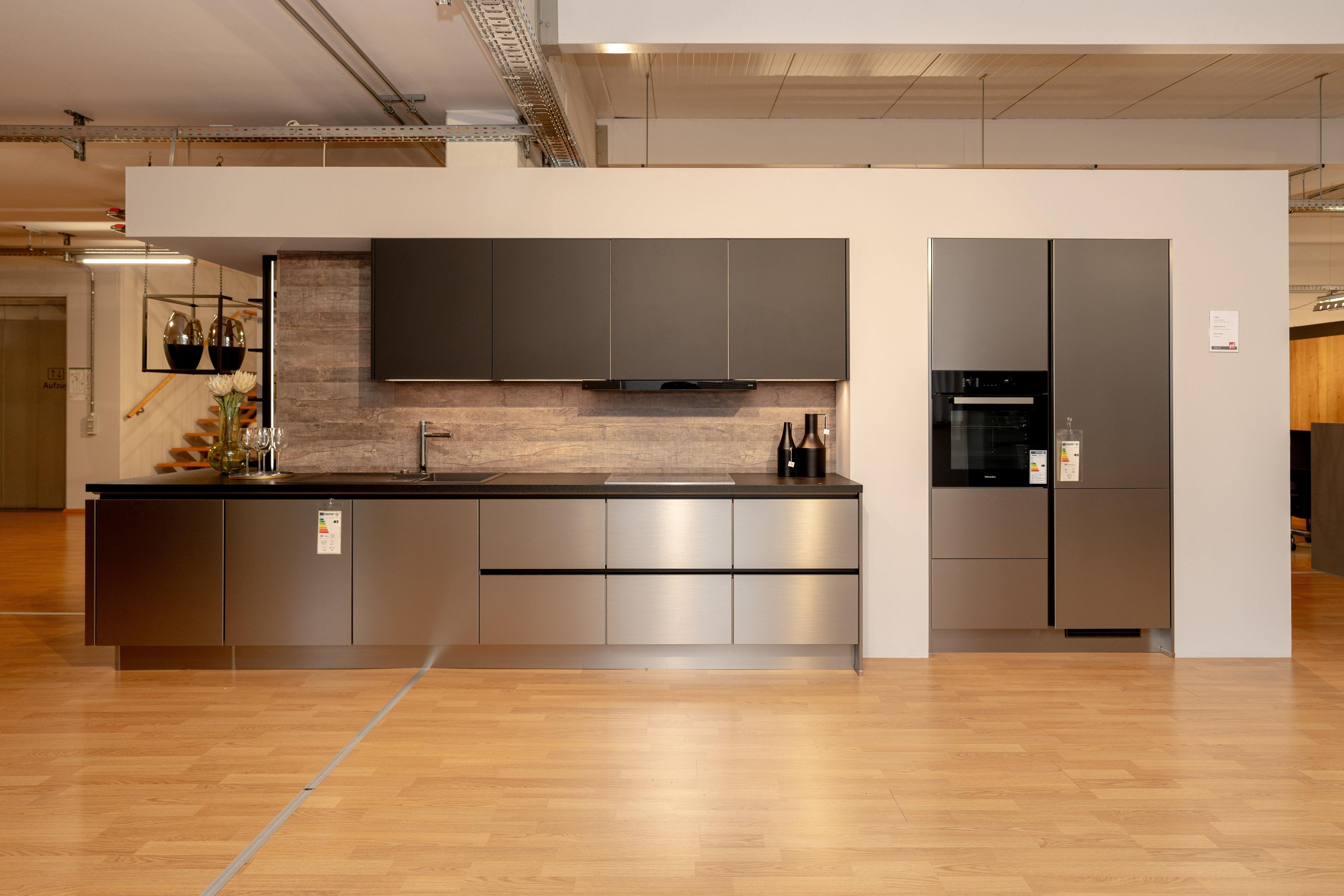 Moderne Küchenzeile mit anthrazitfarbenen Oberschränken und bronzefarbenen Unterschränken, schwarzer Arbeitsplatte und Holzrückwand. Hochschrank rechts in gleicher Optik, Showroom-Ambiente mit Parkett.