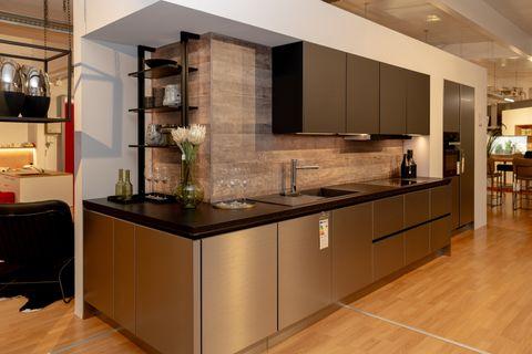 Moderne Küchenzeile mit anthrazitfarbenen Oberschränken und bronzefarbenen Unterschränken, schwarzer Arbeitsplatte und Holzrückwand. Hochschrank rechts in gleicher Optik, Showroom-Ambiente mit Parkett.