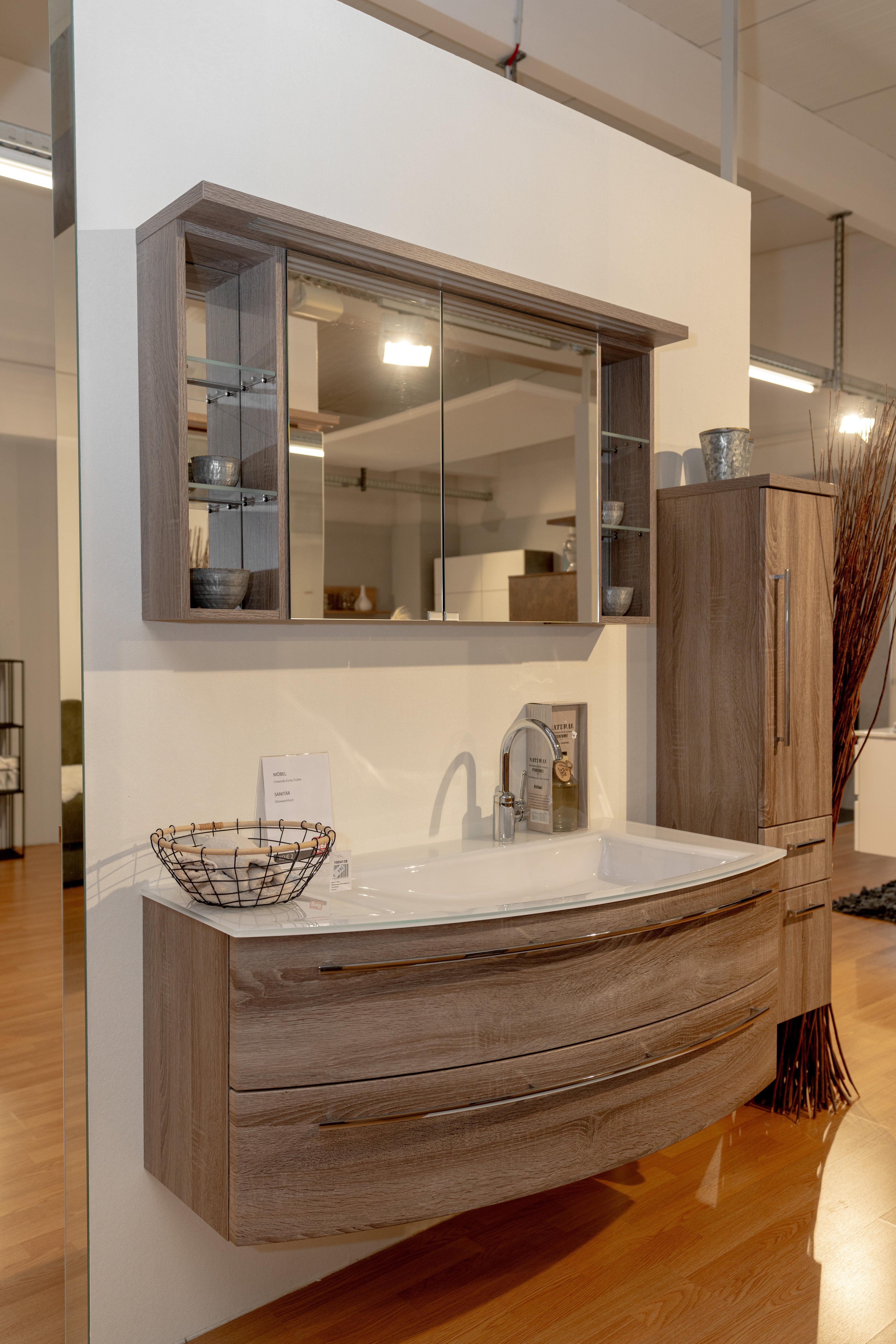 Moderner Waschtischunterschrank in Holzoptik mit weißem Waschbecken, Chromarmatur und dekorativem Drahtkorb in Badausstellung.