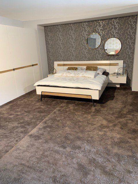 Schlafzimmer-Showroom mit weißem Bett vor ornamentaler grauer Tapete und weißem Eckkleiderschrank mit Holzakzenten.