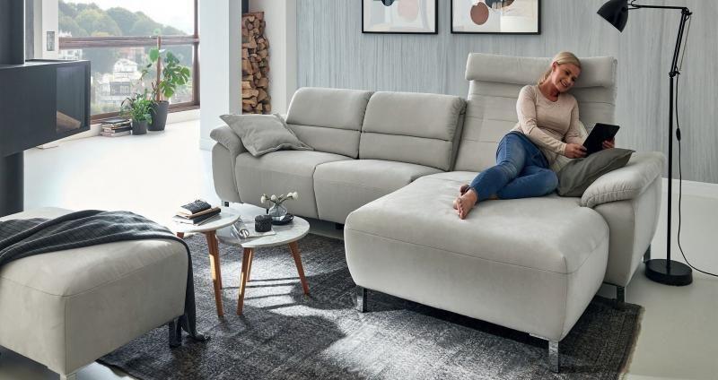 Graue Polstergarnitur mit entspannter Frau auf der Chaiselongue zeigt Wohnkomfort vor Panoramafenstern.
