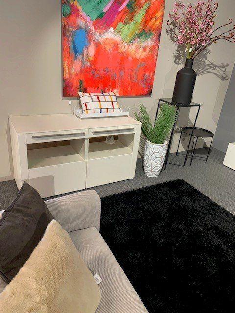 Moderne Wohnzimmereinrichtung mit weißem Sideboard, abstraktem Kunstwerk und dekorativen Pflanzen in geometrischen Übertöpfen.