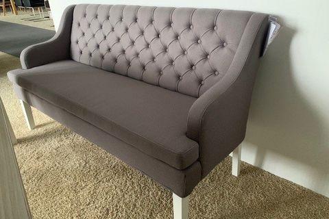 Elegante graue Polsterbank mit Chesterfield-Knopfheftung, Armlehnen und weißen Holzbeinen auf beigem Hochflorteppich.