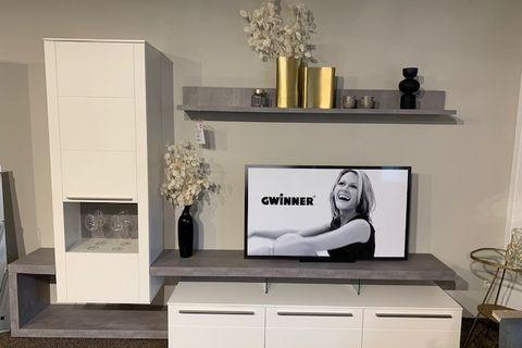 Moderne Wohnwand in Weiß und Grau mit TV, Regalen und schwarzem Relaxsessel vor großen Fenstern.