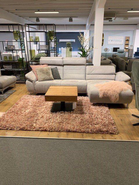 Hellgraue Eckcouch mit Chaiselongue und Holz-Couchtisch auf rosa Hochflor-Teppich in Möbelhaus-Showroom.
