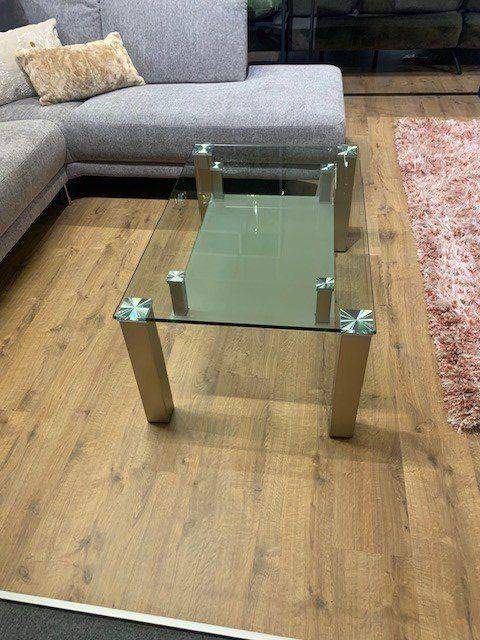 Quadratischer Glas-Couchtisch mit zwei Glasebenen und Edelstahl-Gestell vor modernem Sofa.