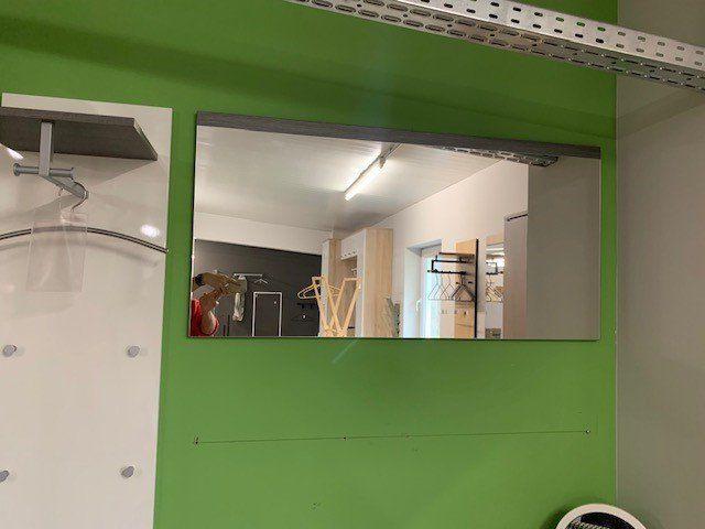 Wandspiegel der Garderobe mit Blick in den dahinterliegenden Ausstellungsraum mit Möbeln und Accessoires.