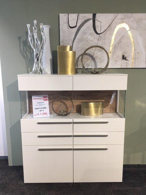 Weißes Sideboard mit Schubladen, Preisschild und modernen Deko-Objekten in Gold und Silber.