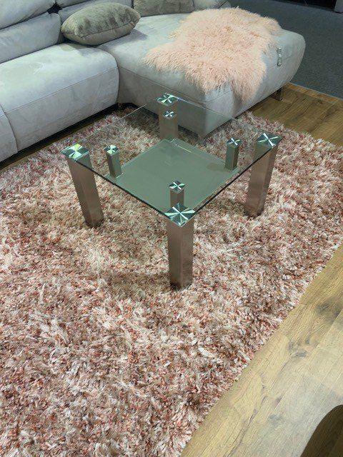 Quadratischer Glas-Couchtisch mit Edelstahlfüßen vor hellem Sofa auf strukturiertem Teppich.