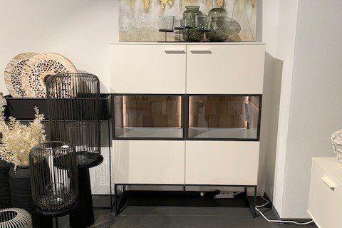 Weißes Highboard mit LED-beleuchteten Glasfächern und Holzrückwand, dekoriert mit Vasen vor großem Fenster