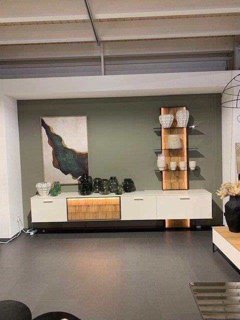 Weißes TV-Lowboard mit Holzakzenten und wandmontiertem Regal vor grüner Wand in Möbelhaus-Ausstellung.