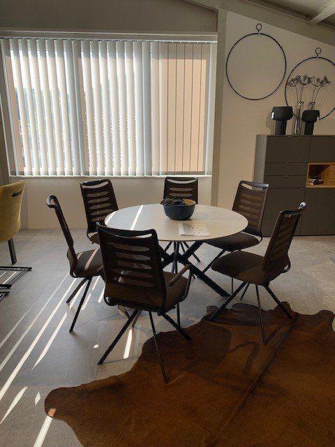 Runder weißer Esstisch umgeben von sechs grauen Stühlen mit Lamellrückenlehne in lichtdurchflutetem Showroom.