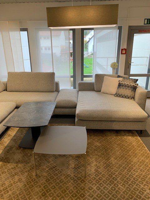 Graue Polstergarnitur mit Chaiselongue und weißem Couchtisch auf beigem Teppich vor großen Fenstern.