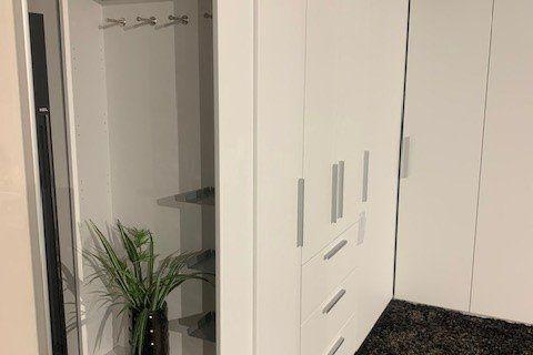 Weißer Eckkleiderschrank mit Schubladen-Mittelteil auf dunklem Teppich unter Dachschräge mit sichtbaren Balken.