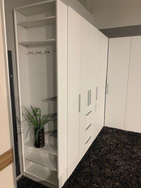 Geöffneter weißer Eckschrank zeigt Innenaufteilung mit Regalbrettern, Kleiderstange und dekorativer Vase.