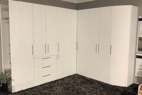 Weißer Eckkleiderschrank mit Schubladen-Mittelteil auf dunklem Teppich unter Dachschräge mit sichtbaren Balken.