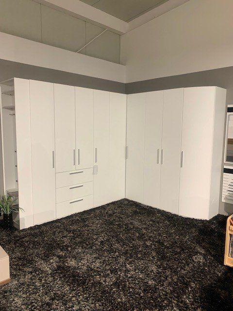 Weißer Eckkleiderschrank mit Schubladen-Mittelteil auf dunklem Teppich unter Dachschräge mit sichtbaren Balken.
