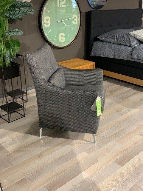 Dunkelgrauer Sessel mit gemustertem Bezug und Metallbeinen neben schwarzem Bett und großer Wanduhr.
