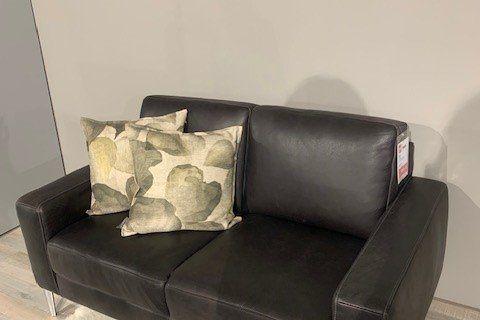 Schwarzes Ledersofa mit grünen Dekokissen auf Metallfüßen vor Kuhfell-Teppich in Möbelhaus-Ausstellung.