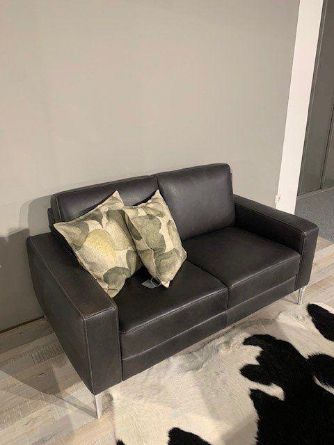 Schwarzes Ledersofa mit grünen Dekokissen auf Metallfüßen vor Kuhfell-Teppich in Möbelhaus-Ausstellung.