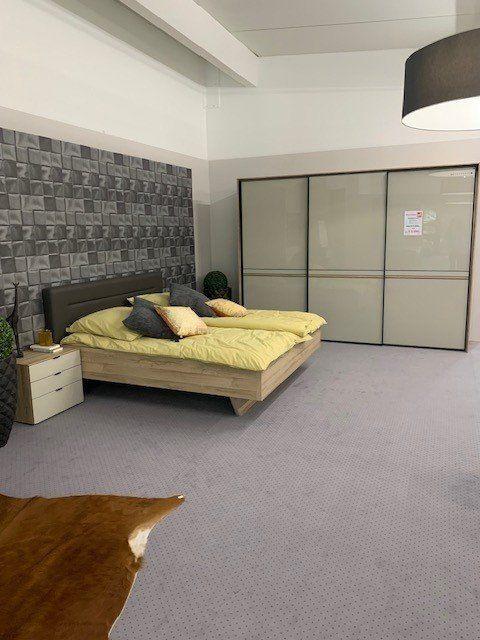 Schlafzimmer-Showroom mit gelber Bettwäsche, weißen Nachttischen und anthrazitfarbenem Schwebetürenschrank unter sichtbaren Deckenbalken.