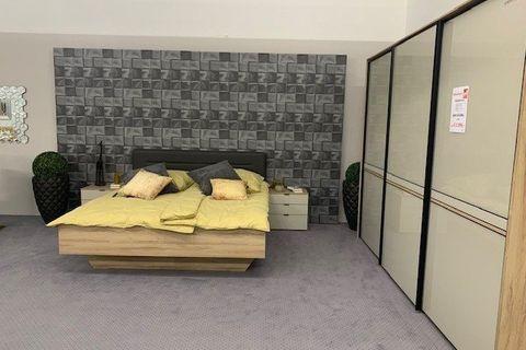 Moderne Schlafzimmerausstellung mit Eichenbett, grauer 3D-Wandverkleidung und großem Schwebetürenschrank mit Glasfronten.
