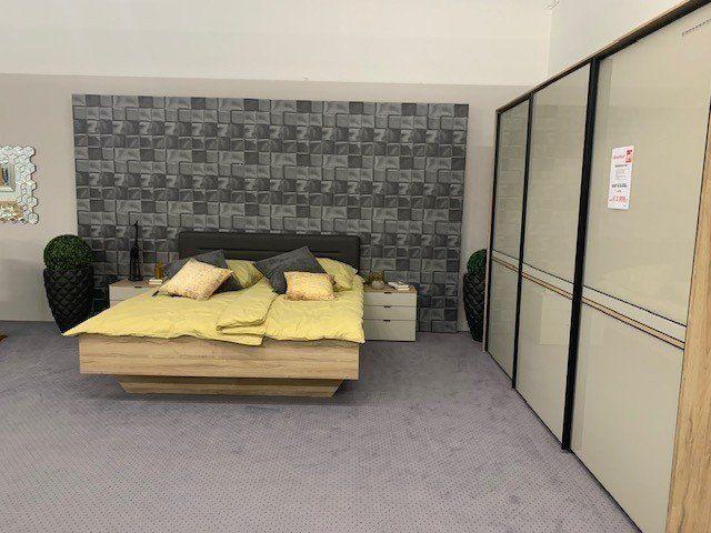 Moderne Schlafzimmerausstellung mit Eichenbett, grauer 3D-Wandverkleidung und großem Schwebetürenschrank mit Glasfronten.