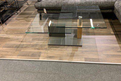 Couchtisch aus Glas mit goldenem Metallgestell vor grauem Sofa in Möbelhaus-Ausstellung.