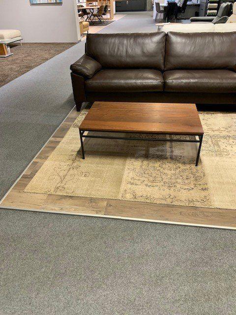 Couchtisch aus Holz mit schwarzem Metallgestell vor grauem Ledersofa auf beigem Teppich in Möbelhaus-Showroom.