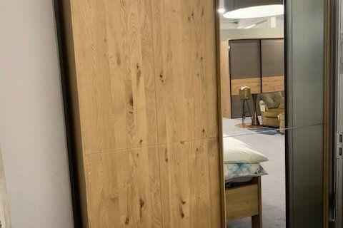 Moderner Kleiderschrank aus hellem Eichenholz mit Spiegeltür, teilweise geöffnet mit Blick ins Schlafzimmer.