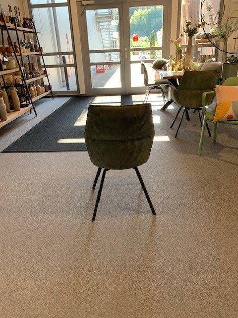Rückansicht des olivgrünen Cord-Stuhls in sonnendurchflutetem Showroom mit modernen Möbeln im Hintergrund.