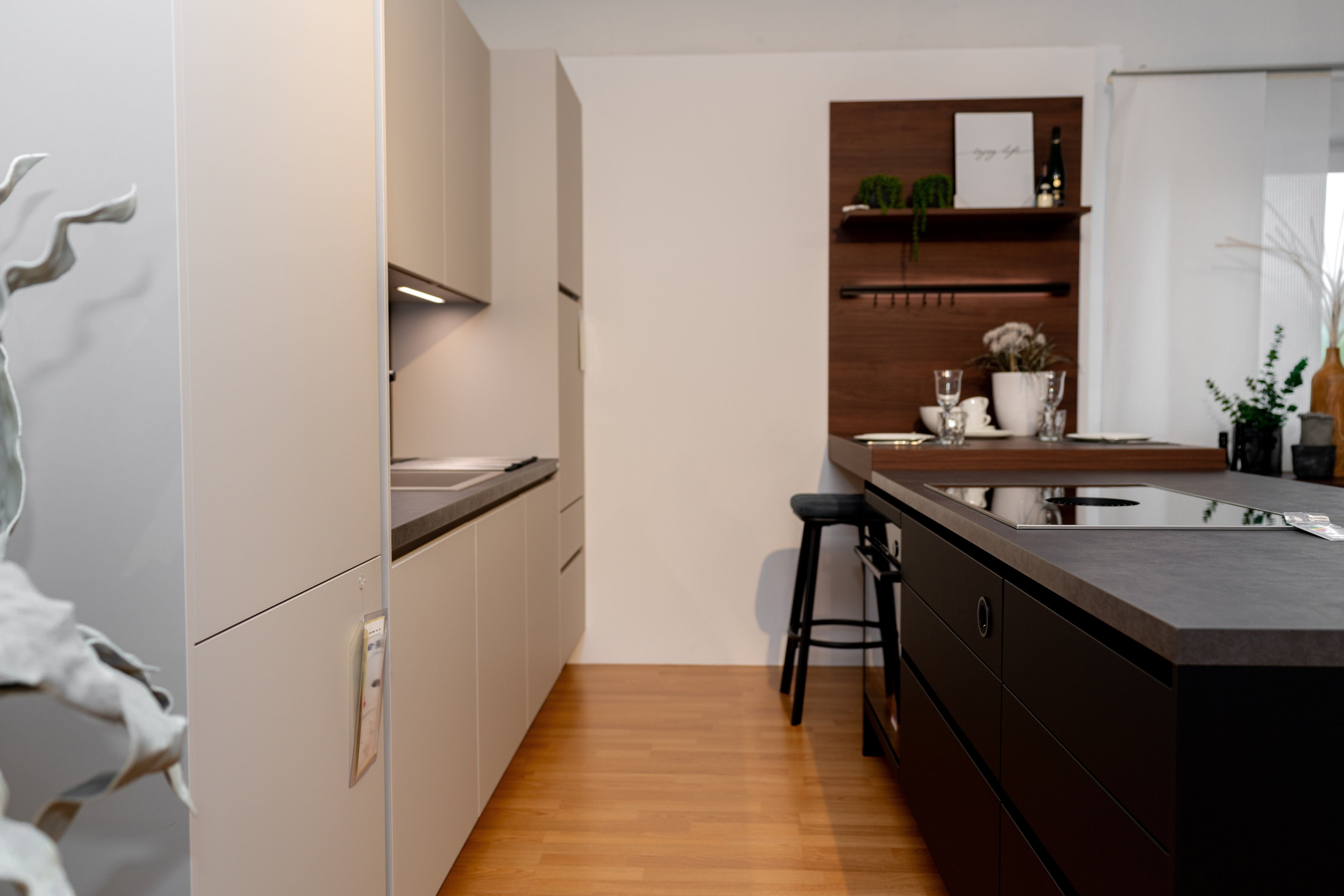 Moderne Küche mit beigen Oberschränken, schwarzer Kücheninsel mit grauer Arbeitsplatte und Nussbaum-Wandregal mit Pflanzen und Gläsern. Schwarze Barhocker stehen vor der Kücheninsel.
